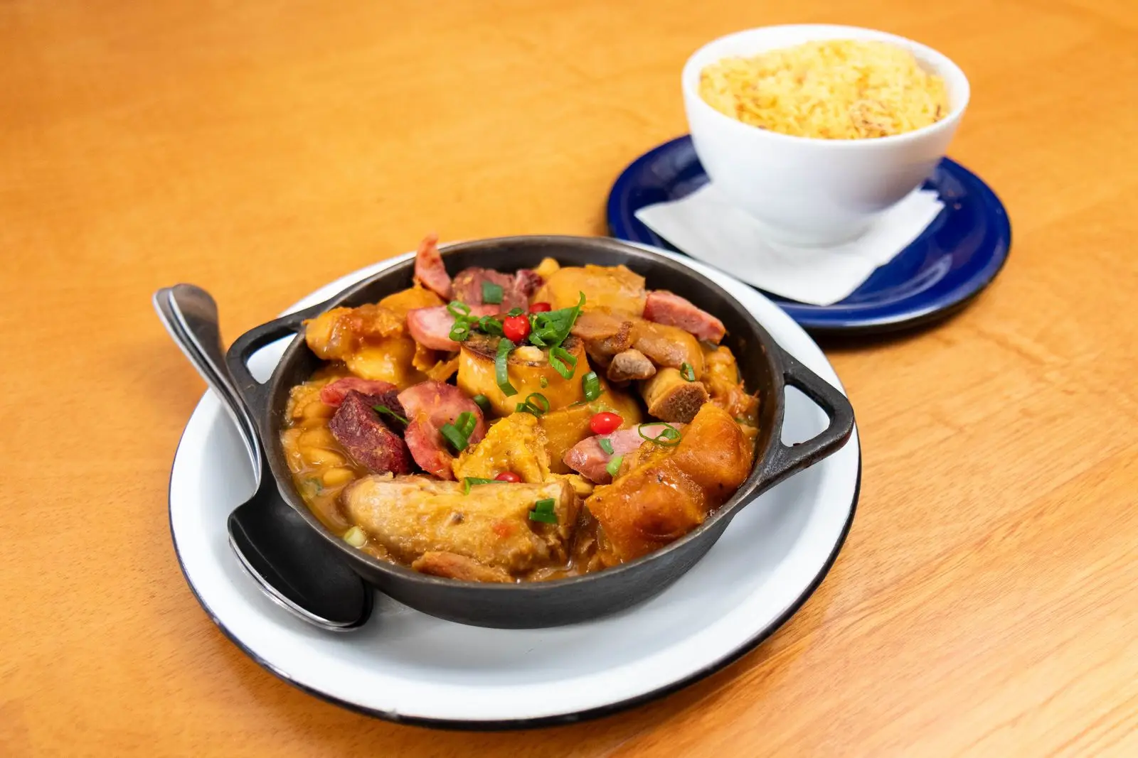 Panela de ferro com linguiça, carne e legumes ensopados, ao lado de um pote de farofa